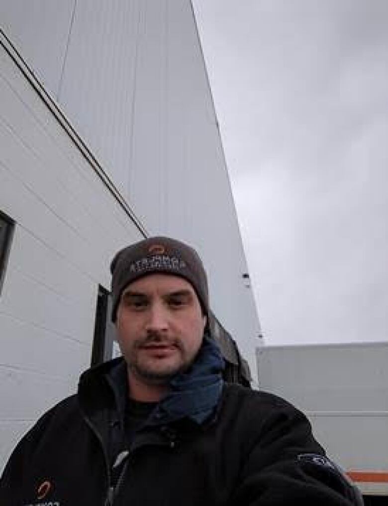 A man with a grey beanie and facial hair poses for a selfie. He appears to be outside, near a large white building. 