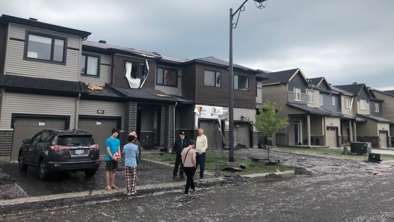 People stand outside homes with damaged exteriors and hail on the ground.