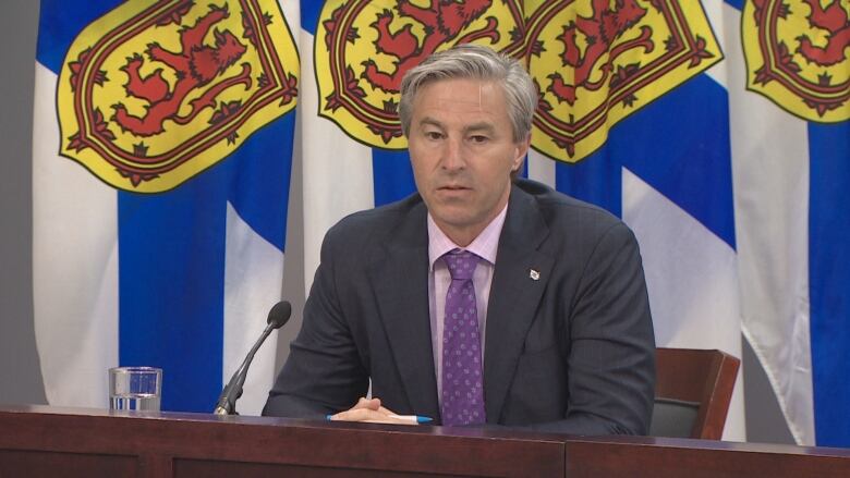 A man wearing a suit and purple tie sits at a desk with Nova Scotia flags behind it.