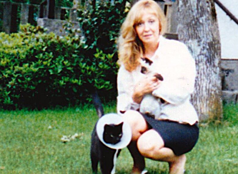 A woman with blonde hair, a white shirt and black skirt kneels with a black cat on a green lawn.