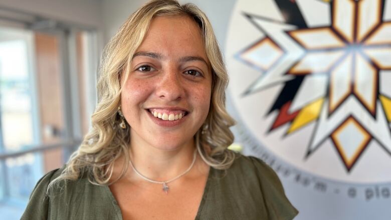 Shelby Arsenault-Ellands, a smiling woman with blond hair, stands in front of a Mi'kmaq emblem painted on a wall. 