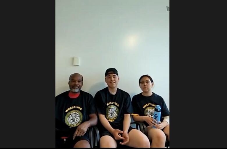 A man sits next to two teenagers, all wearing black t-shirts that say wrestling. 