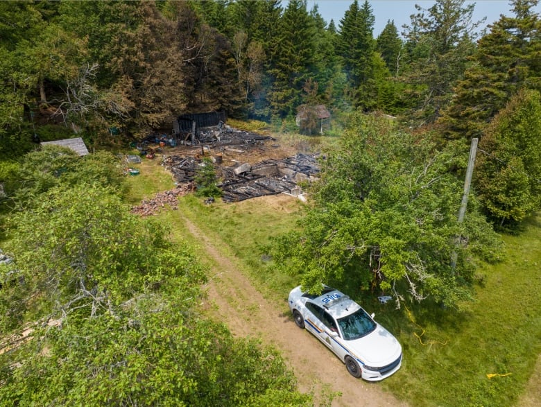 A drone shot of an RCMP cruiser parked on a grassy area in front of a burned-down building.