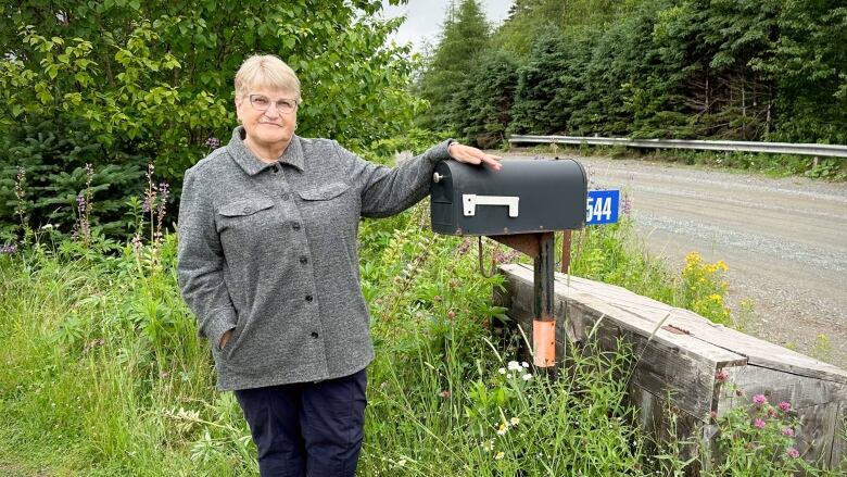 A woman with glasses stands beside the mailbox at the end of her driveway next to a gravel road.