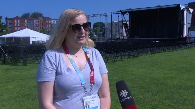 A woman with a colourful NAIG lanyard is standing on the grass speaking into a microphone. Tents, chairs, and a large stage are set up behind her.