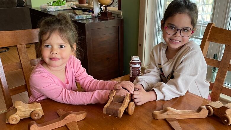 Two young girls smile as they sit at a wooden table and hold a wooden hippo toy. There are other wooden toys on the table.