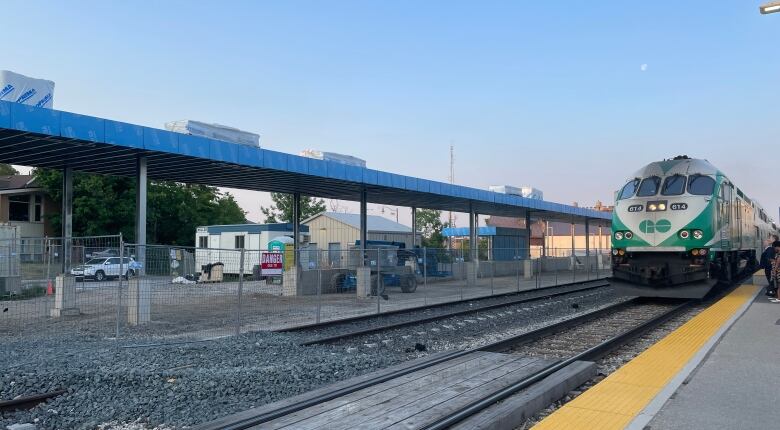 Green and white train on tracks is in right part of photo, construction with concrete pillars can be seen in the background.