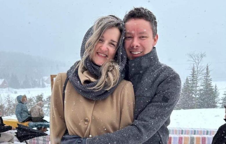 A man hugs a woman as they both smile at the camera. Snow falls around them.