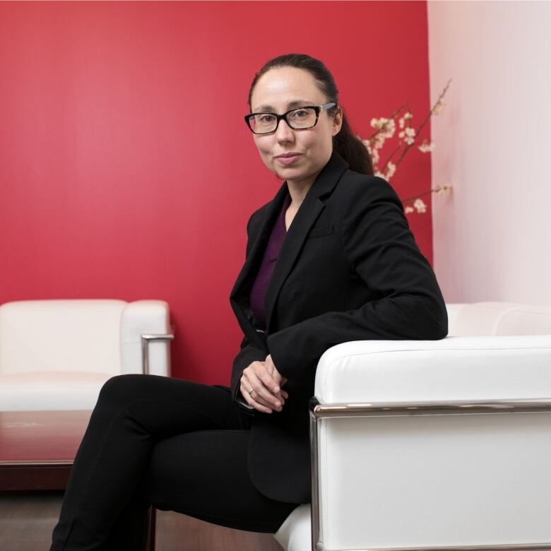 A woman wearing glasses and a black suit, with her hair in a ponytail, smiles as she sits on a white chair, posing for a portrait in front of a red wall.