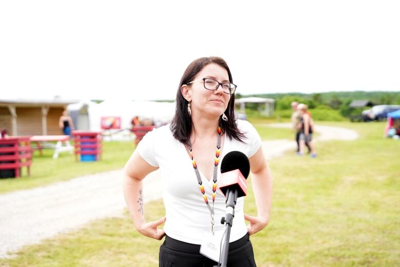 A woman with brown hair and glasses stands in front of a microphone. Behind her are people walking, tents with vendors inside them and picnic tables.