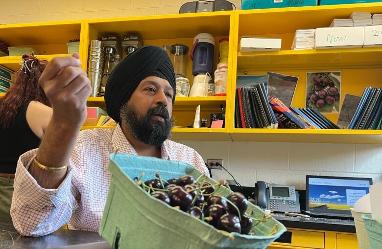 A man with beard and a black turban speaks in a room with a box of cherries sitting beside him .