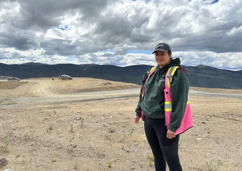 A woman in a safety vest stands on a rocky field with trucks and mountains in the distant background.