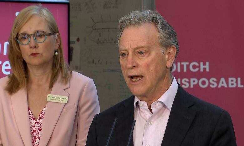 A man in a light pink shirt and suit jacket speaks from a rostrum as a woman with blond, shoulder-length hair stands to his right.