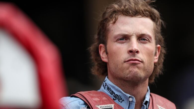 A man looks at the camera, wearing a vest, with a mullet.