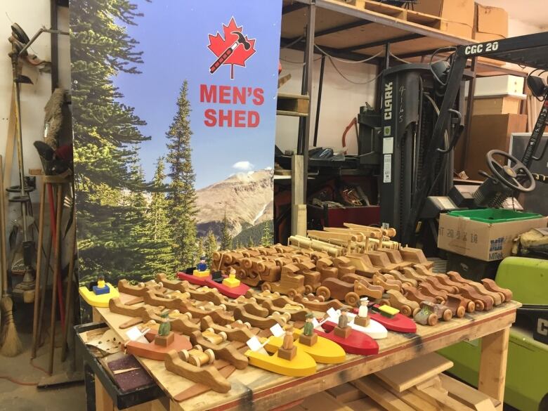 A collection of wooden toy boats, cars and planes sits on a table with a banner for Men's Shed in the background.