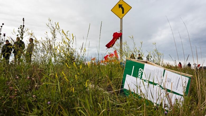 A wooden placard bearing paper notices lies on its side in a ditch alongside a road.