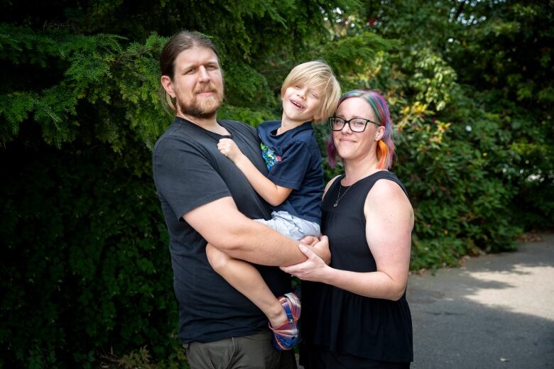 A man holds a blonde boy in his hands, with a woman with coloured hair gripping his palms.