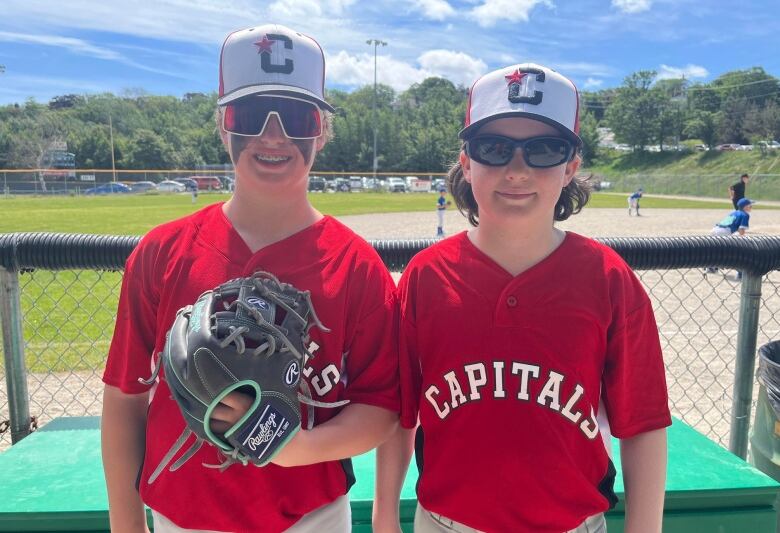 Two boys stand in front of a baseball field wearing red Capitals jersey's. One is wearing a baseball mitt. 