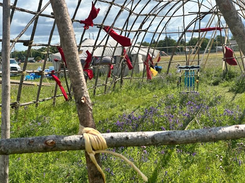 A wooden chair sits in the grass inside a bare wooden structure. A tent sits in the background.