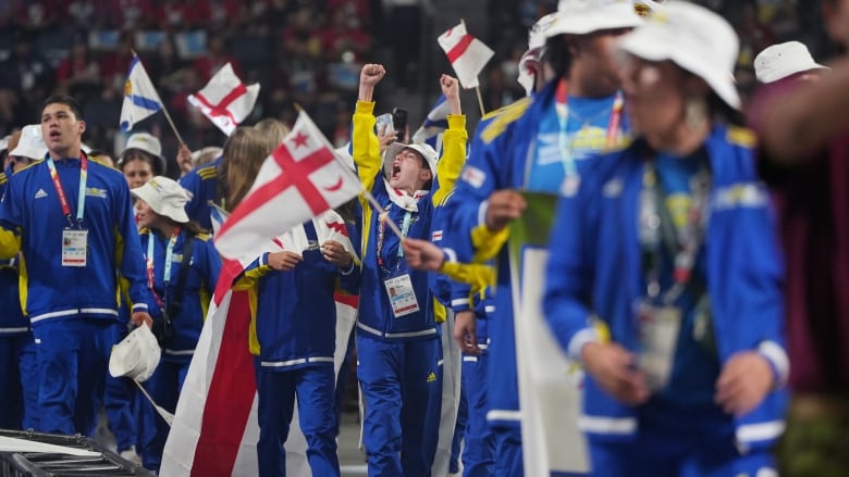 athletes wearing blue and yellow track suits march with red and white flags. A boy in the centre has his arms raised and his mouth wide open/