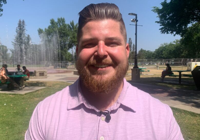 A smiling man with a beard stands in a park with a fountain behind him. 