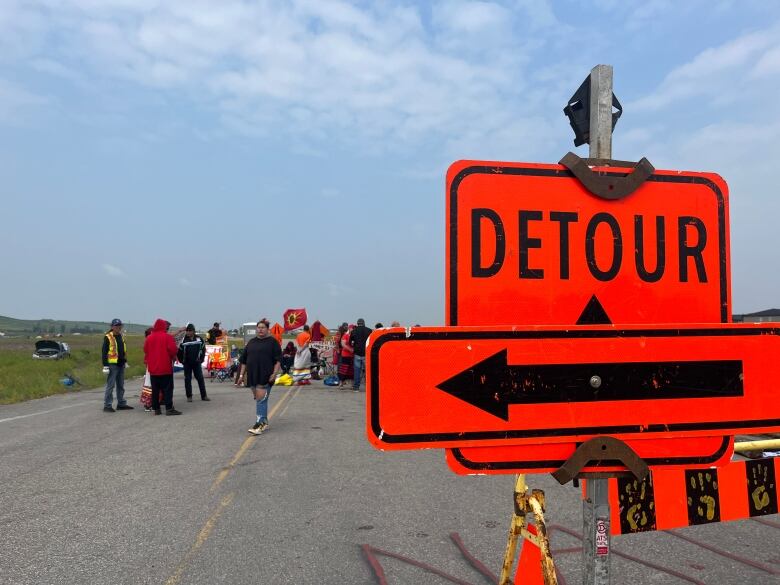 An orange sign with the word, Detour, on it is seen in the foreground. In the background is a crowd of people around a blockade.