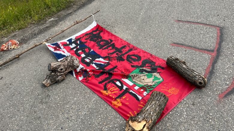 A Manitoba provincial flag lies on pavement, held down by three pieces of wood. The phrase Heartless Heather is painted across the flag.