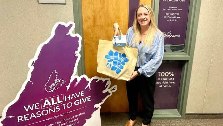 A woman smiles for a photo while holding a bag with flowers on it. She stands next to a sign that says, 'We all have reasons to give.'