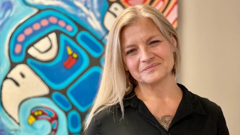 A blonde woman in a black shirt poses in front of a colourful piece of Indigenous artwork. 