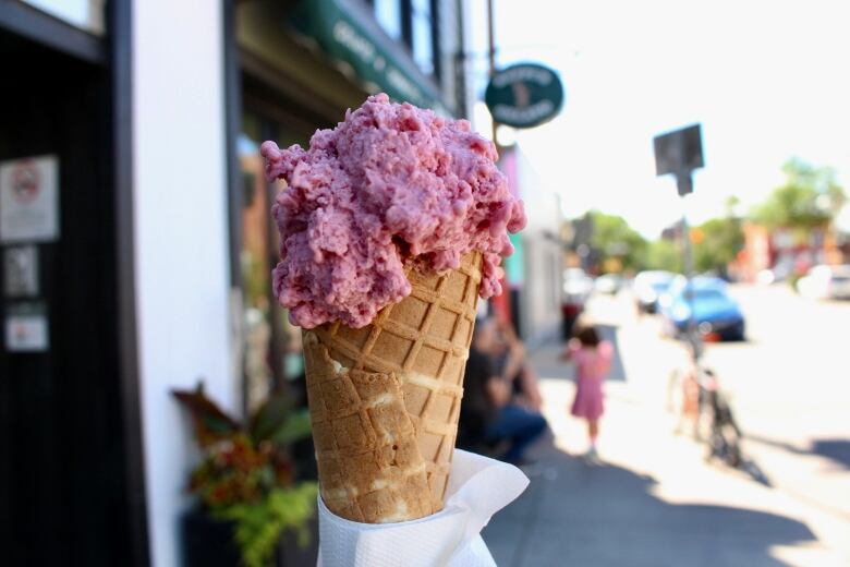 A close-up shot of a waffle cone topped with a pink ice cream being held on a commercial street with a business in the background.