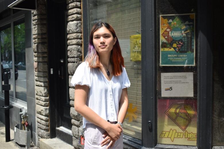 A person wearing a white top and light grey pants stands outside a building. 