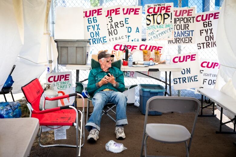 A man sits among signs which read 'CUPE On Strike' in an outdoor tent.