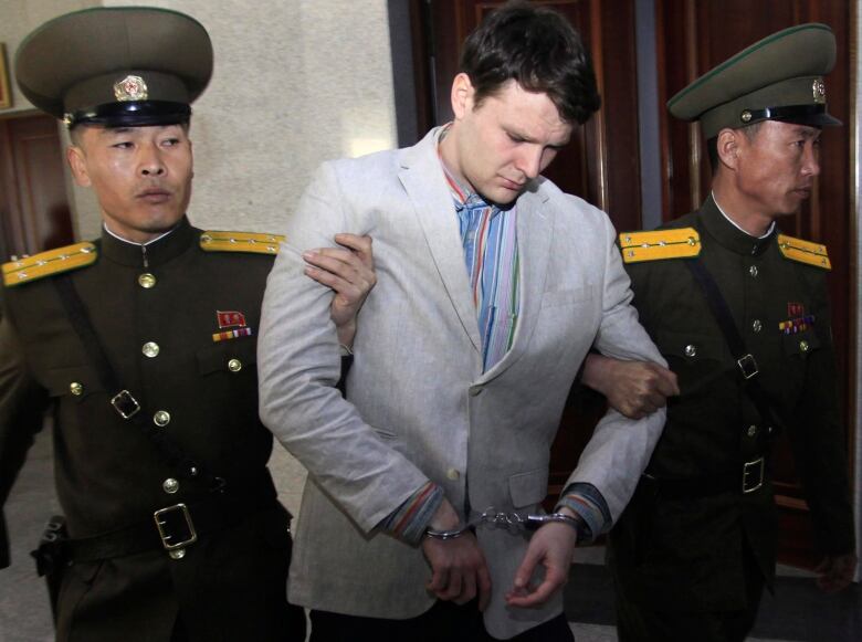 A man in a suit jacket looks downward as he is handcuffed and escorted by two men wearing military caps and uniforms.