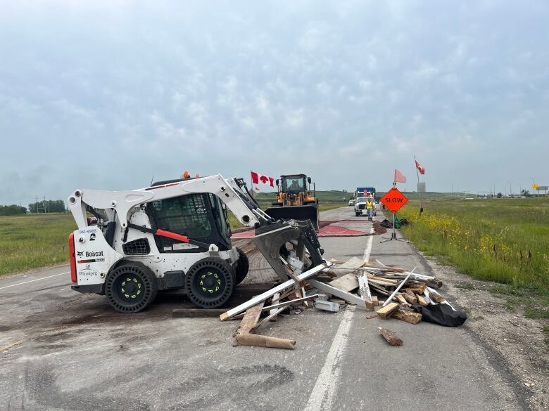 A skid steer breaks apart pieces of wood. An upside down Canada flag is seen in the background.