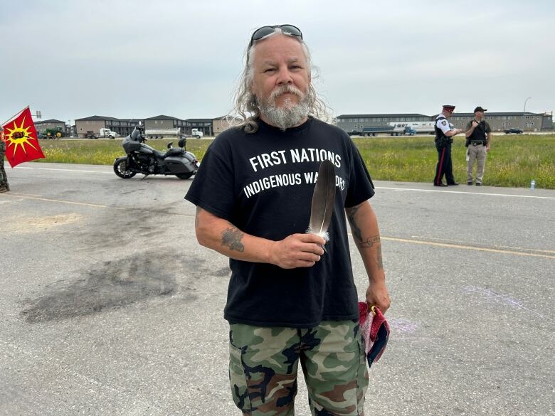 A man in camo pants, black shirt and long white hair holds an eagle feather.