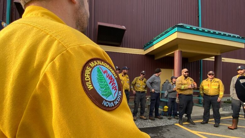 A group of people are seen standing outside a building, with someone's arm seen in the foreground with a badge reading, 'Wildfire Management Nova Scotia.' 