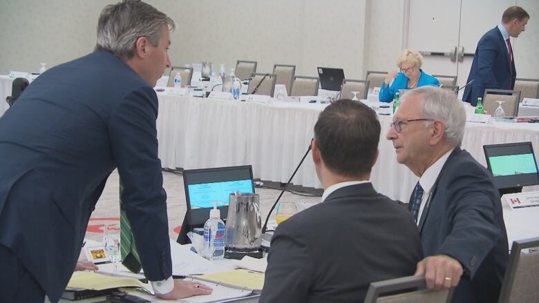 Two men sit at a table with their backs to the camera. A third man leans on the table speaking with them.