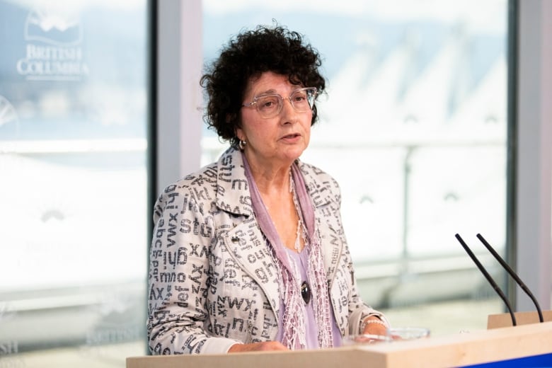 A white woman with curly grey hair speaks in front of a lectern.