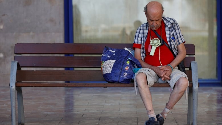 A man sits on a bench with a small fan hanging around his neck with a bag resting beside him. 