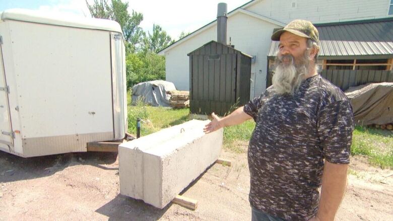 A man with short grey hair and a long grey beard wearing a dark green baseball hat and grey patterned tee-shirt and jeans motions to his right, where a large rectangular cement block sits on the ground in front of the hitch of a commercial trailer.