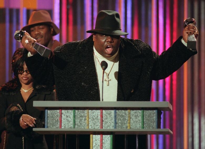 A man in a coat and hat holds out awards in each hand with arms extended while at a podium onstage.