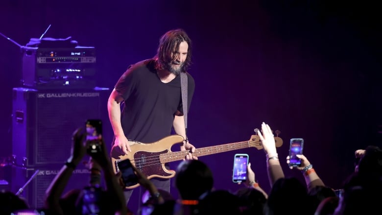 Keanu Reeves holding his bass onstage for a concert while wearing a black T-shirt and smiling at a crowd.