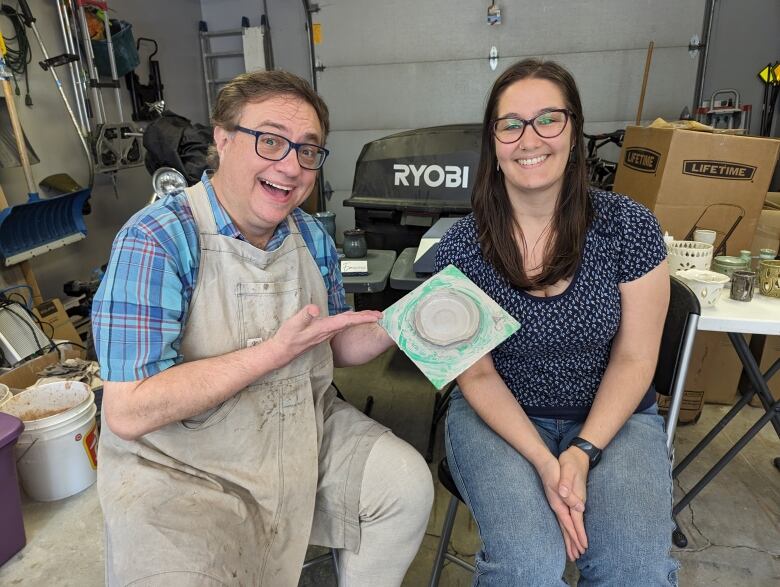 A man smiles while holding a clay bowl. A woman smiles sitting beside him. 