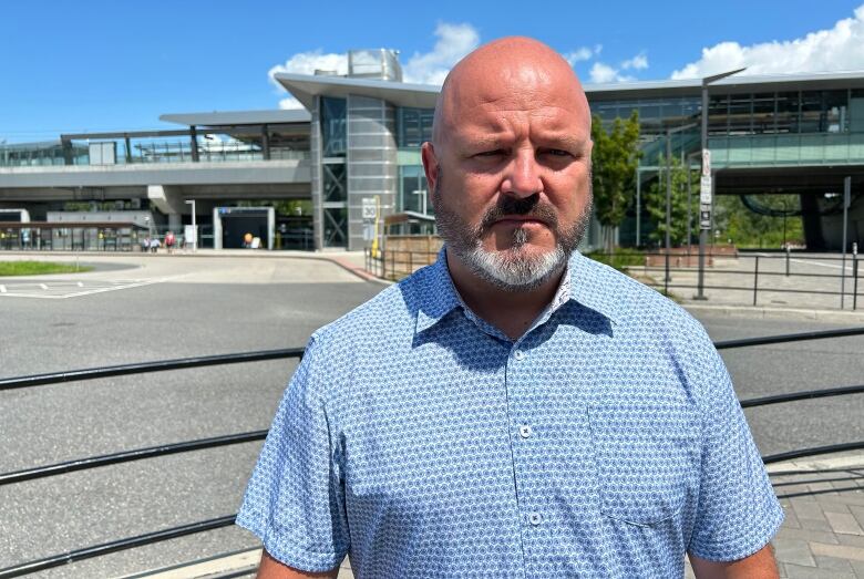 Man in a blue shirt looks straight into a camera without smiling, outside an empty transit station.