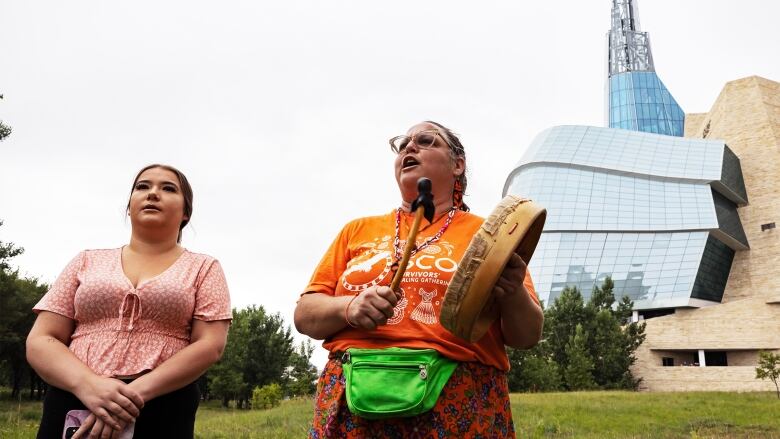 Two women stand on a lawn in front of a museum. One wears an orange shirt and plays a drum. 