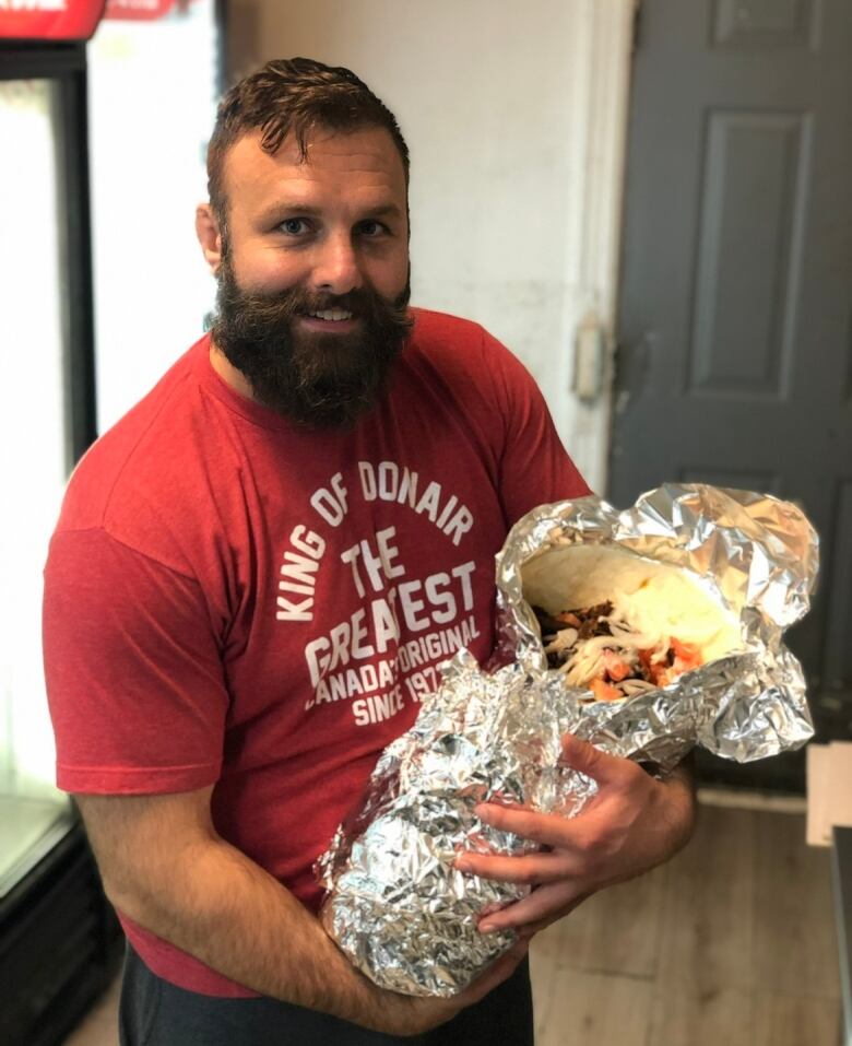 A man with a beard, wearing a red T-shirt, cradles a massive donair in his arms.