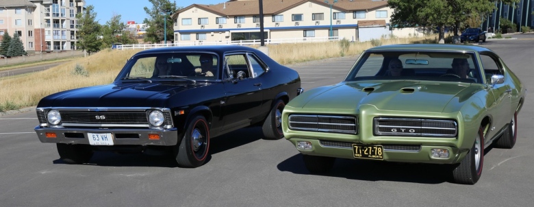 Two restored classic cars on a street. A black 1969 Nova L78 and a light lime green 1969 GTO RAIII.