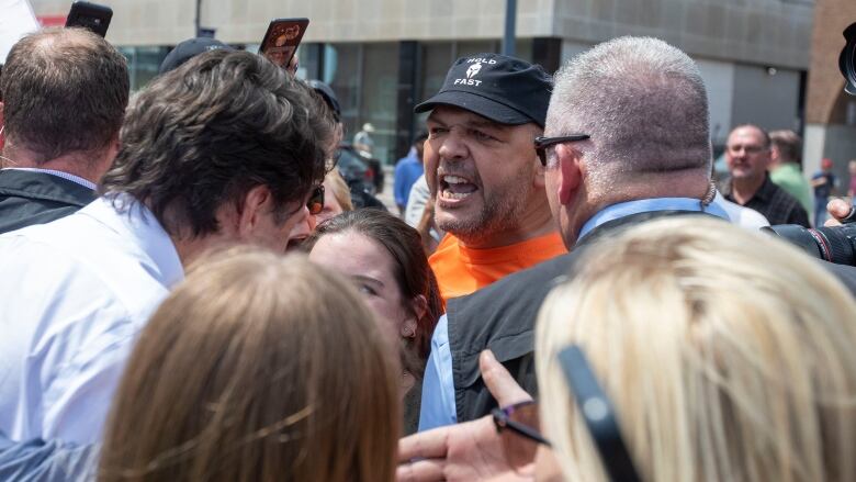 A man wearing a baseball cap shouts at Prime Minister Justin Trudeau as he shakes hands in a crowd outside.