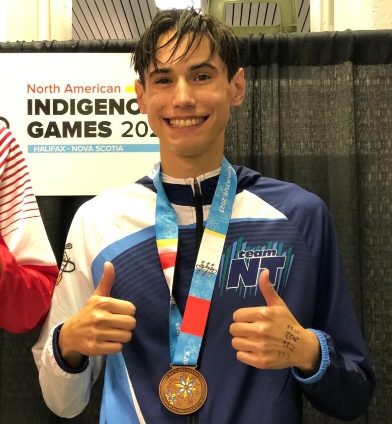 A smiling young man wearing a medal gives the thumbs-up.