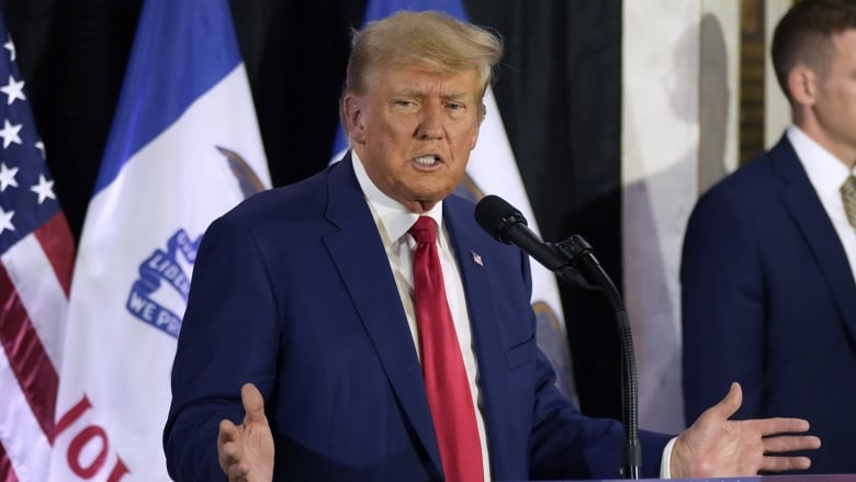 A man in a suit gestures with his hands at the podium in front of a backdrop of flags.